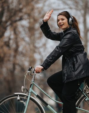 Stylish and confident young businesswoman enjoying a bike ride in the park, waving hello while wearing headphones.